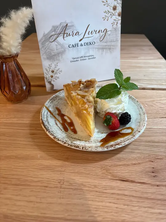 Stück Apfelkuchen mit Mandelblättchen, dazu Sahne, Minze, Erdbeere, Brombeere und Karamellsauce, dahinter die Karte von Aura Living