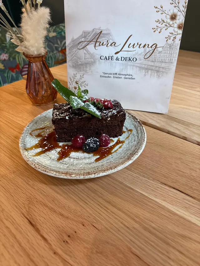 Brownie mit Minze, Erdbeere, Himbeeren, Brombeere, Puderzucker und Karamellsauce auf einem gesprenkelten Teller, dahinter die Karte von Aura Living