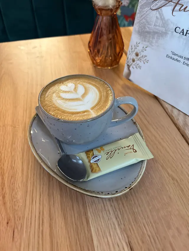 Cappuccino mit Blattmuster im Milchschaum in grau gesprenkelter Tasse, auf der Untertasse Löffel und ein verpackter Keks
