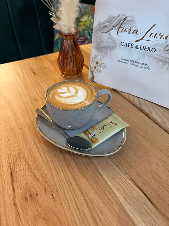 Cappuccino mit Milchschaum-Muster auf grauer Untertasse mit Löffel und Keks, schräg von oben fotografiert