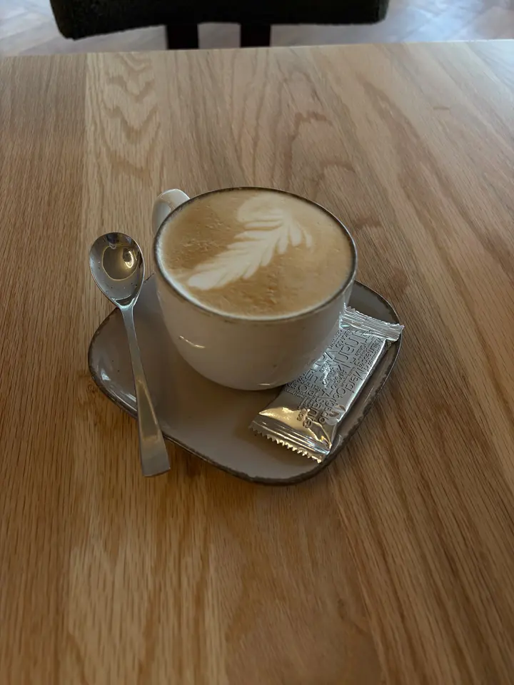 Cappuccino mit Blattmuster im Milchschaum in heller Tasse, auf der Untertasse Löffel und ein verpackter Keks