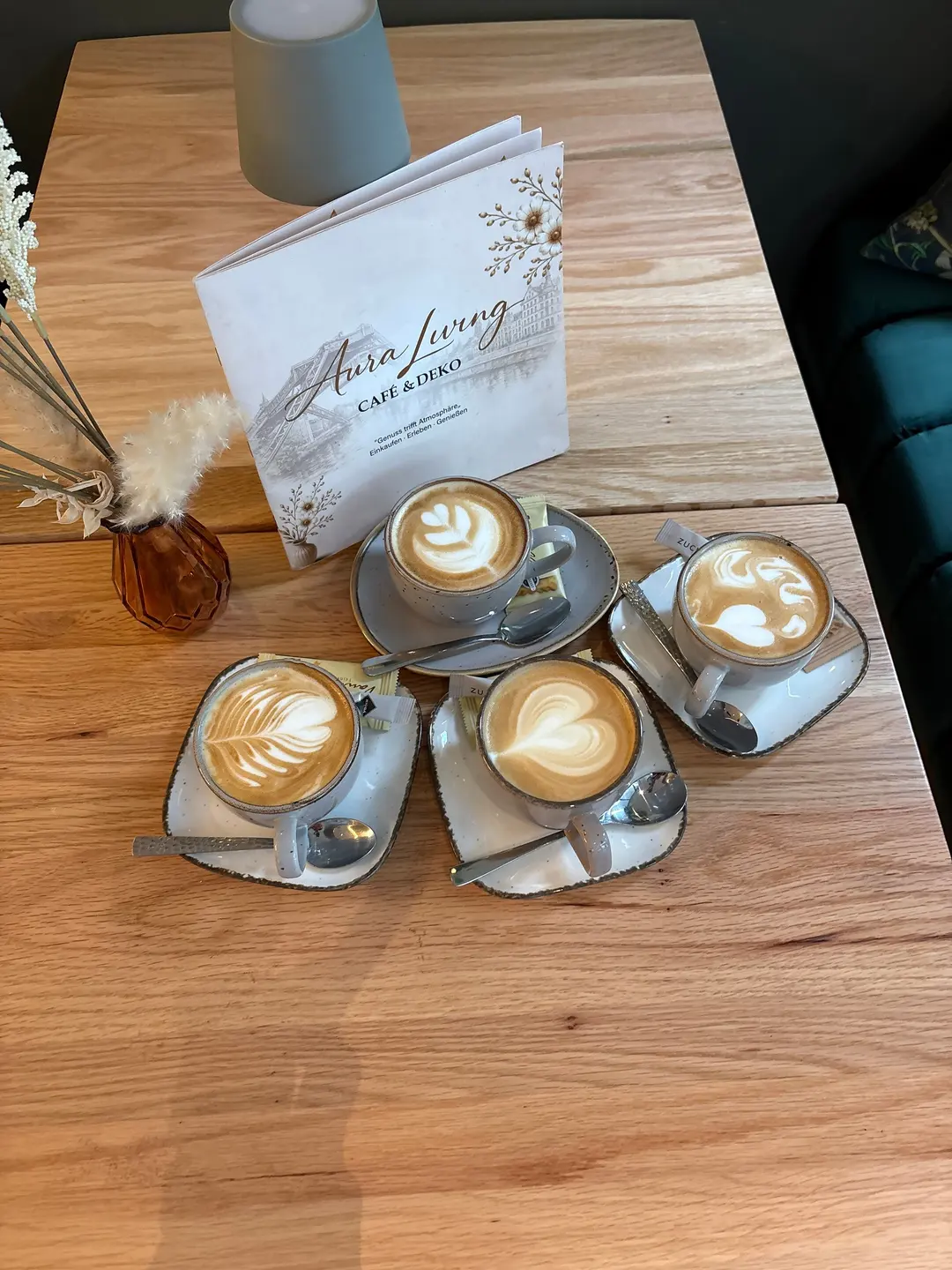 Vier Cappuccino mit Herz- und Blattmustern im Milchschaum in grau gesprenkelten Tassen, dahinter die Karte von Aura Living