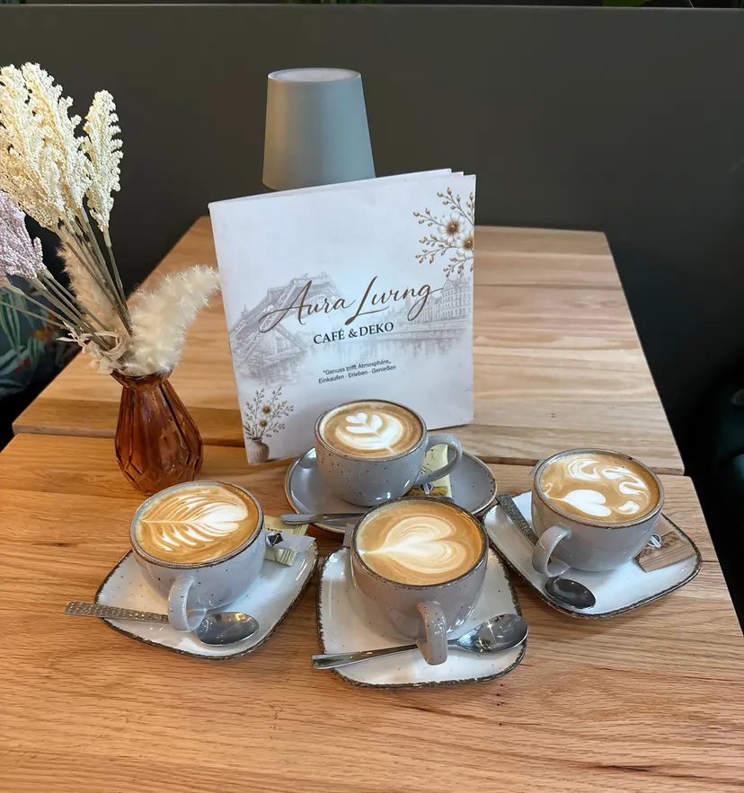 Vier Tassen Cappuccino mit Latte Art auf einem Holztisch, daneben Vase mit Pampasgras und die Karte von Aura Living