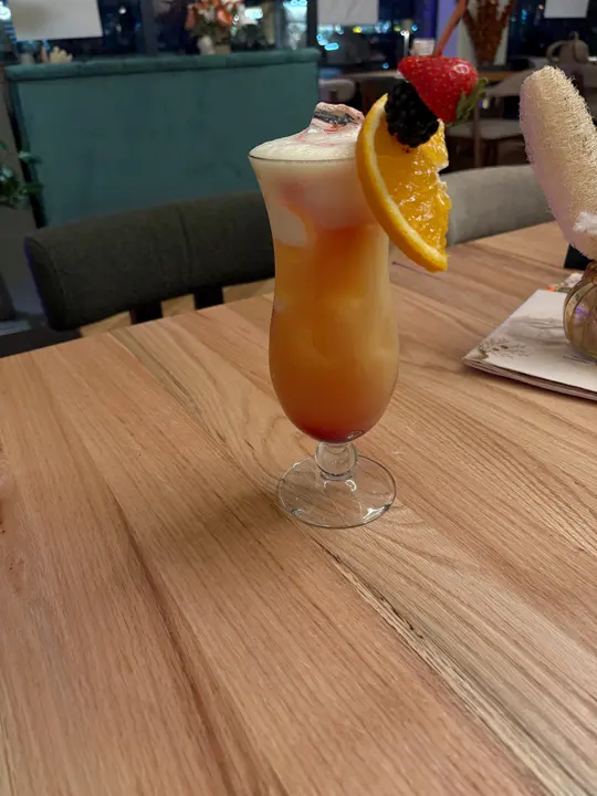 Orange-roter Cocktail im Hurricane-Glas mit Schaumkrone, Orangenscheibe, Brombeere und Erdbeere, am Abend im Gastraum vor einer grünen Polsterbank