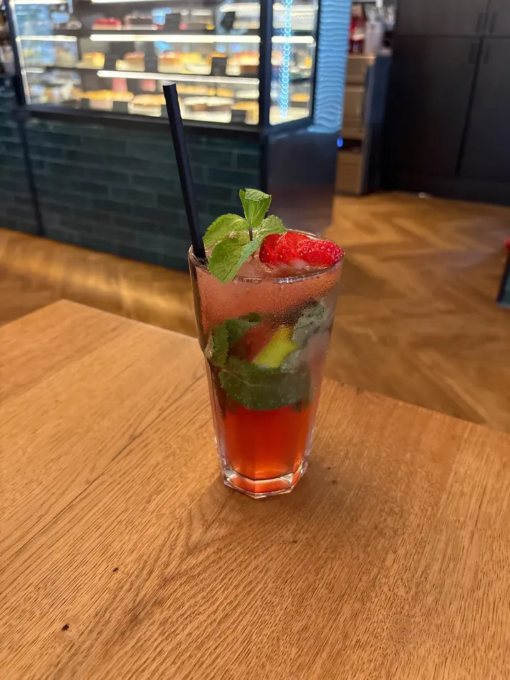 Erdbeer Mojito im hohen Glas mit Minze, Limette und Erdbeere, im Hintergrund die Kuchenvitrine