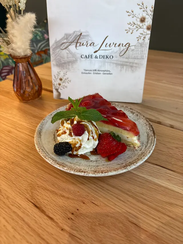 Stück Erdbeerkuchen mit Sahne, Karamellsauce, Minze, aufgefächerter Erdbeere und Brombeere, dahinter die Karte von Aura Living