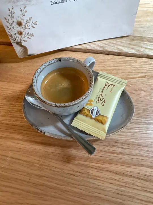 Nahaufnahme einer Tasse Kaffee mit Crema, Löffel und verpacktem Keks auf grauer Untertasse