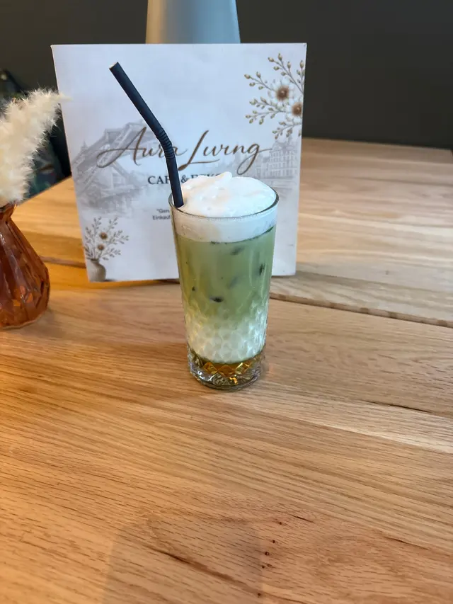 Iced Matcha Latte im geschliffenen Glas mit grüner Matcha-Schicht, Milch, Schaumhaube und schwarzem Strohhalm