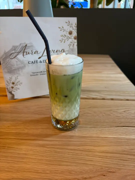 Iced Matcha Latte mit Eiswürfeln und Milchschaum im hohen Glas vor der Karte von Aura Living