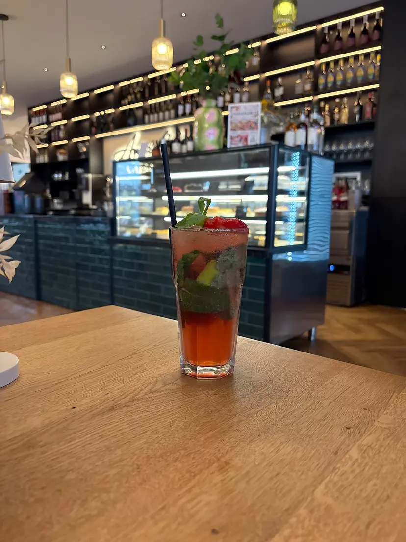 Blick auf die Bar von Aura Living mit beleuchteten Flaschenregalen, grün gefliester Theke und Kuchenvitrine, im Vordergrund ein Erdbeer Mojito