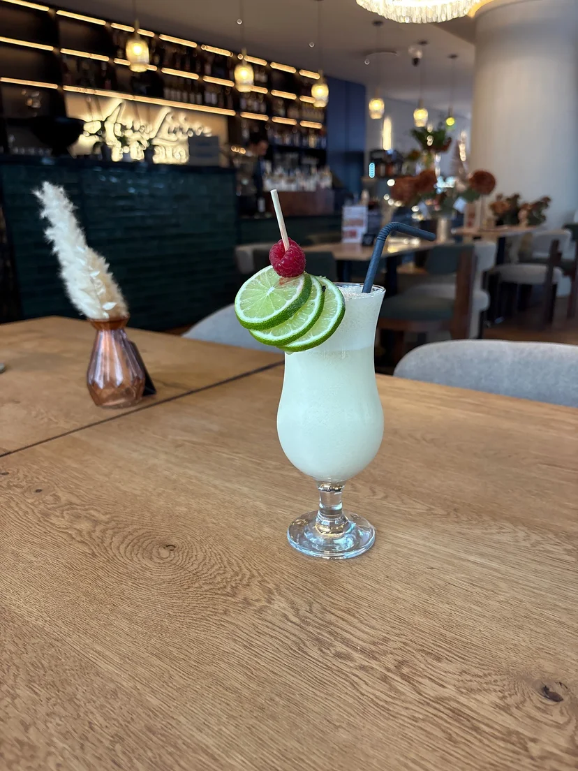 Bar mit beleuchtetem Aura-Living-Schriftzug, Pendelleuchten und Kronleuchter, im Vordergrund eine Pina Colada