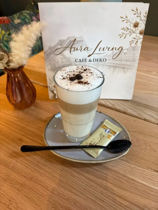 Latte Macchiato im Glas mit drei Schichten, Milchschaum mit Kaffeebohnen und Kakaopulver, auf der Untertasse Löffel und Keks
