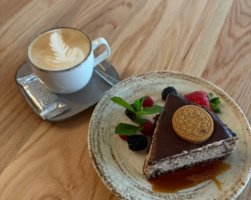 Tortenstück mit Schokoladenglasur, hellem Keks und Karamellsauce neben einem Cappuccino mit Milchschaum-Muster, von oben fotografiert