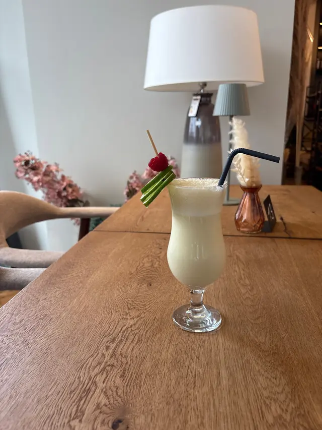 Pina Colada mit Limettenscheiben, Himbeere und Strohhalm auf einem Holztisch vor heller Wand und Tischleuchte