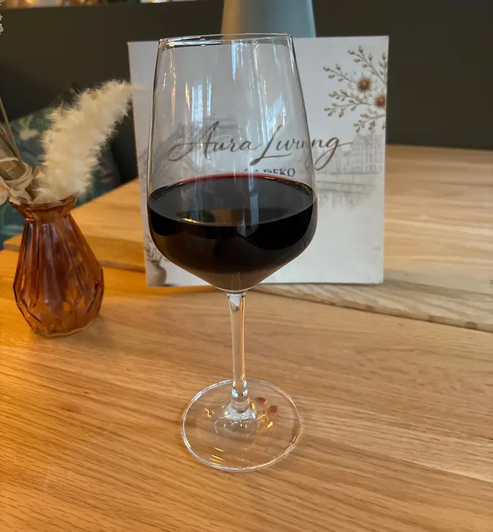 Glas Rotwein auf einem Holztisch, daneben Vase mit Pampasgras und die Karte von Aura Living