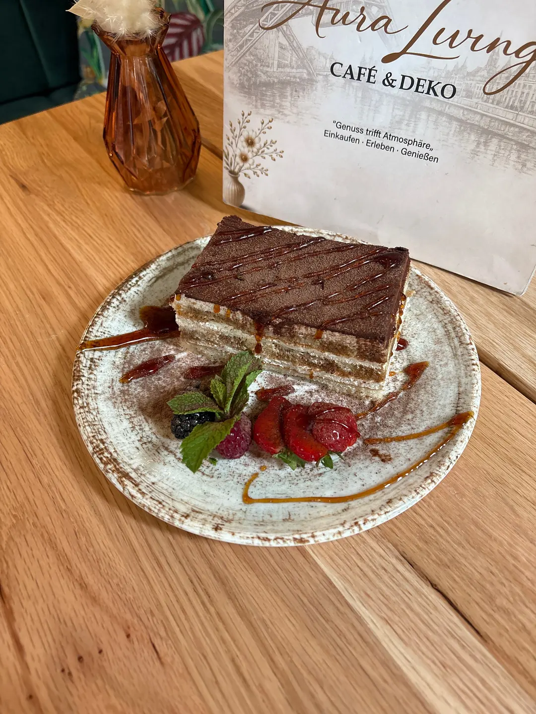 Rechteckiges Stück Schichttorte mit heller Creme und Kakaodecke, garniert mit Schokosauce, Erdbeere, Himbeeren, Brombeere und Minze